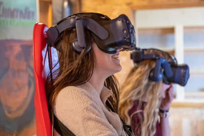 Two women smile while wearing virtual reality headsets in a cozy setting.